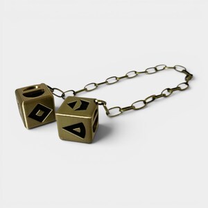 May include: Two brass-colored dice connected by a chain. Each die has black geometric symbols on its faces. The dice are cube-shaped and appear to be made of metal. The chain is also brass-colored and links the two dice together.