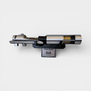 May include: A silver lightsaber prop, displayed on a black stand. The stand features a Star Wars logo and a nameplate that reads "GALEN MAREK Lightsaber". The lightsaber has a detailed design with a black grip and clear sections.