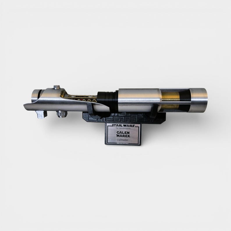 May include: A silver lightsaber prop, displayed on a black stand. The stand features a Star Wars logo and a nameplate that reads "GALEN MAREK Lightsaber". The lightsaber has a detailed design with a black grip and clear sections.