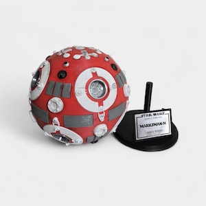 May include: A red and white spherical Star Wars Marksman-H training remote prop. The prop has a detailed design with gray and black accents. It sits on a black stand with a silver nameplate that reads "STAR WARS Episode IV: A New Hope" and "MARKSMAN-H - Training Remote".