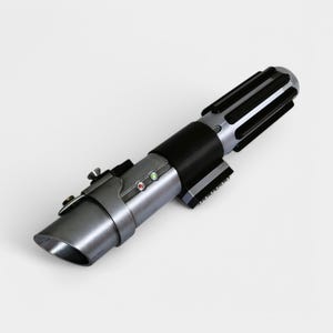 May include: A silver and black lightsaber with a black handle and a silver blade. The lightsaber has a small, black button on the side.