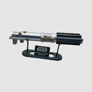 May include: A silver and black metal lightsaber replica featuring a textured grip, gold-colored accents, and a circular orange button. The lightsaber rests on a black display stand with a plaque that reads STAR WARS Anakin Skywalker Lightsaber.