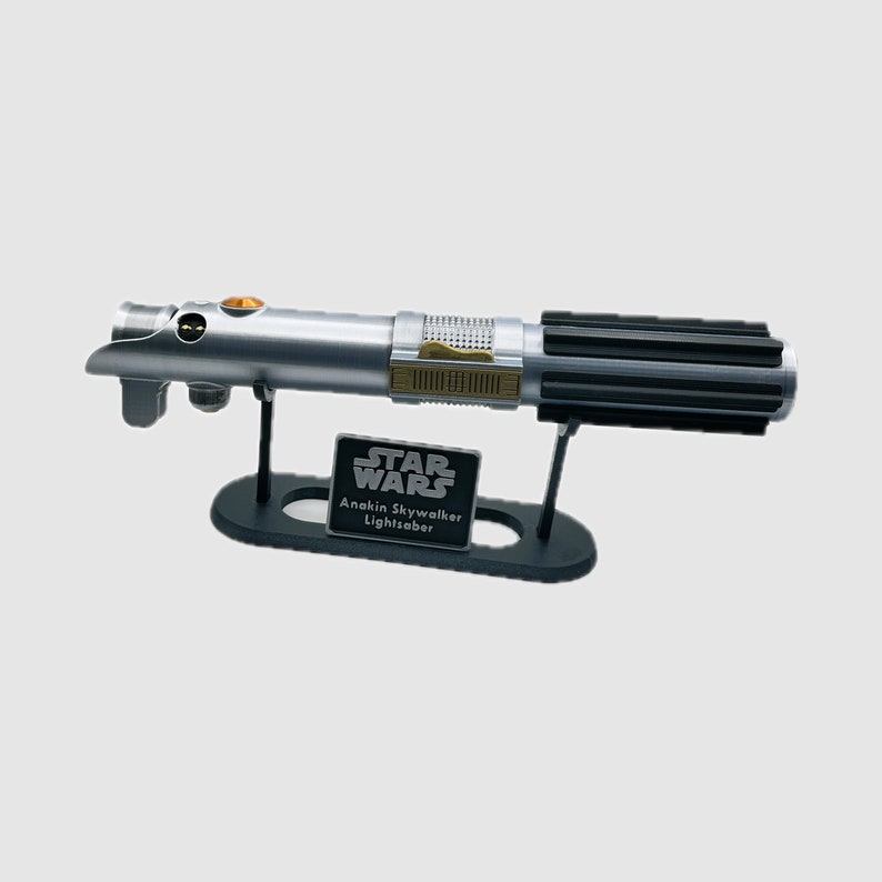 May include: A silver and black metal lightsaber replica featuring a textured grip, gold-colored accents, and a circular orange button. The lightsaber rests on a black display stand with a plaque that reads STAR WARS Anakin Skywalker Lightsaber.