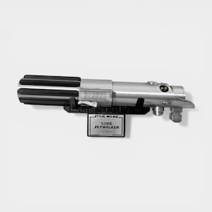May include: A silver and black lightsaber prop, with a black base and a plaque that reads "STAR WARS, Luke Skywalker, Lightsaber". The lightsaber has a detailed design with a ribbed handle and metallic finish.