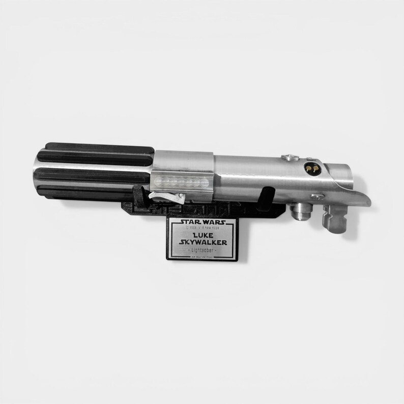 May include: A silver and black lightsaber prop, with a black base and a plaque that reads "STAR WARS, Luke Skywalker, Lightsaber". The lightsaber has a detailed design with a ribbed handle and metallic finish.