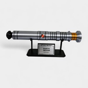 May include: A silver lightsaber prop with a black handle and gold accents, displayed on a black stand. The prop has a red button and is labeled "Darth Revan - Lightsaber" with the Star Wars logo.