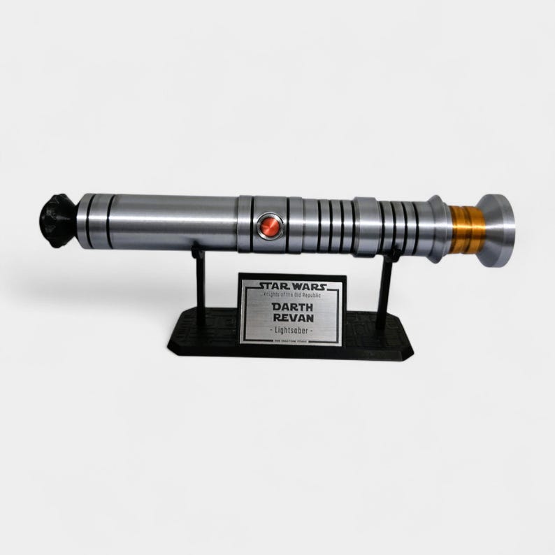 May include: A silver lightsaber prop with a black handle and gold accents, displayed on a black stand. The prop has a red button and is labeled "Darth Revan - Lightsaber" with the Star Wars logo.