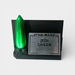May include: A black display stand featuring a vibrant green Kyber Crystal. The stand includes a silver plaque with the text "STAR WARS JEDI GREEN - Kyber Crystal - ". The crystal is illuminated, creating a striking visual effect.