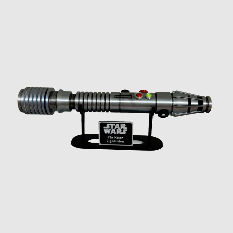 May include: A silver Star Wars lightsaber with a green blade, on a black stand. The stand has a plaque that reads "Star Wars, Plo Koon Lightsaber".