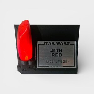 May include: A black display stand featuring a glowing red Kyber Crystal, with the text "STAR WARS" and "SITH RED - Kyber Crystal -" on a silver plaque. The stand is a collectible item.