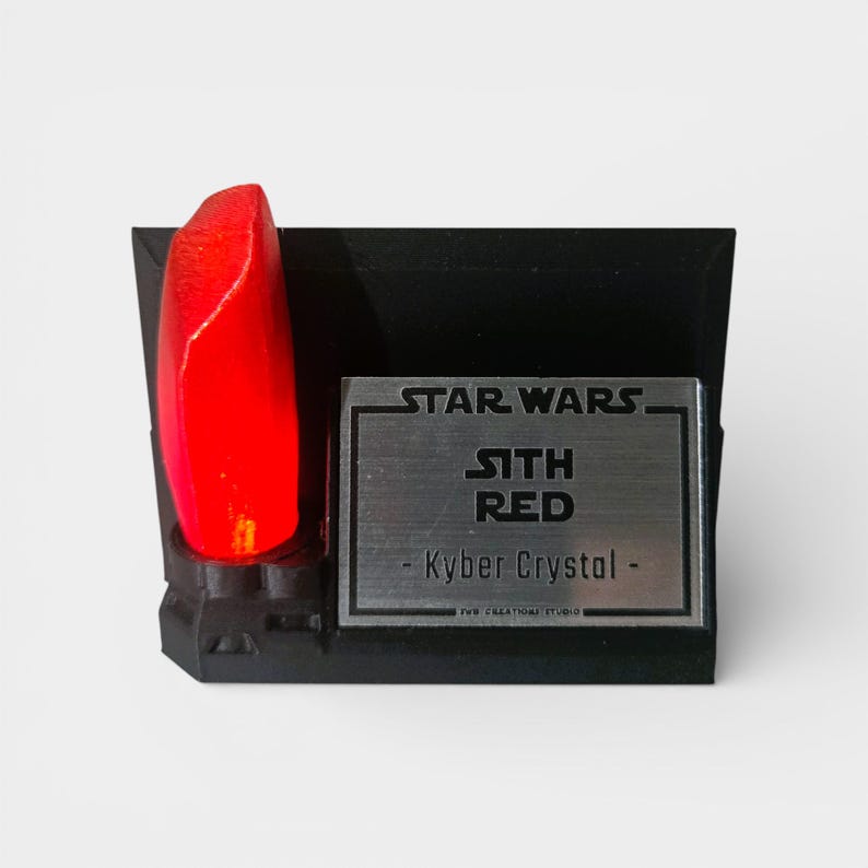 May include: A black display stand featuring a glowing red Kyber Crystal, with the text "STAR WARS" and "SITH RED - Kyber Crystal -" on a silver plaque. The stand is a collectible item.