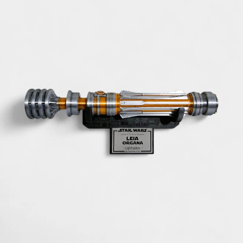 Puede incluir: Un sable de luz de la franquicia Star Wars, con un dise&ntilde;o met&aacute;lico en plata, dorado y negro. Est&aacute; montado en un soporte negro con una placa con el texto "Leia Organa - Lightsaber".
