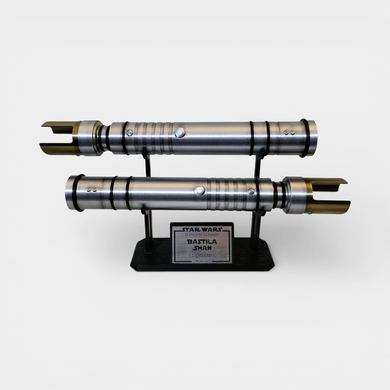 May include: Two silver lightsabers with black accents and gold blades are displayed on a black stand. The lightsabers are labeled "Bastila Shan" from Star Wars. The lightsabers are approximately 12 inches long.
