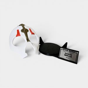 May include: A white Darth Nihilus mask with red and gold detailing, beside a black stand and a nameplate. The nameplate reads "STAR WARS Knights of the Old Republic II DARTH NIHILUS - Mask".