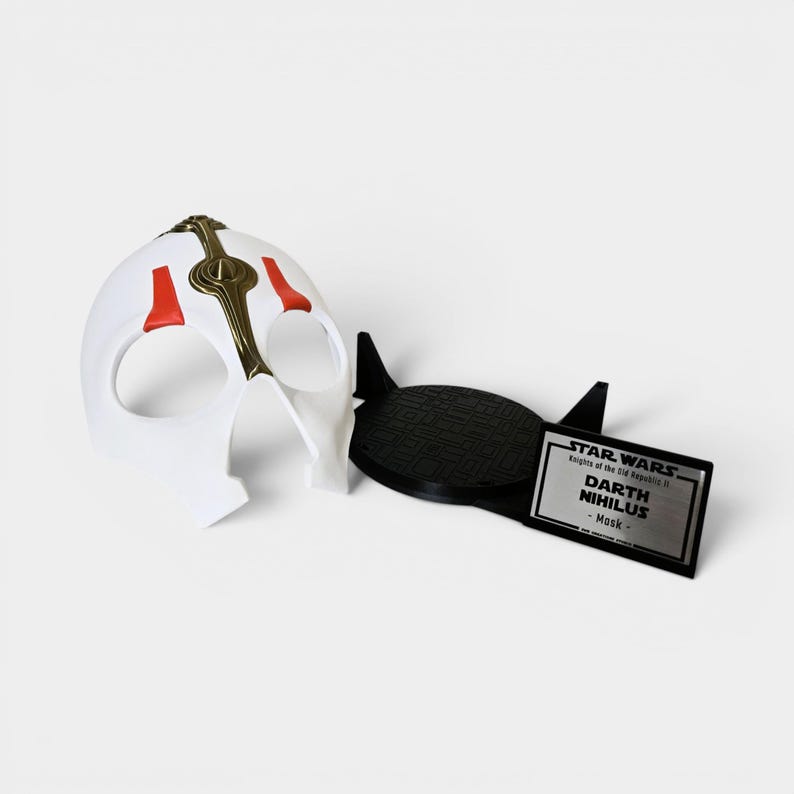 May include: A white Darth Nihilus mask with red and gold detailing, beside a black stand and a nameplate. The nameplate reads "STAR WARS Knights of the Old Republic II DARTH NIHILUS - Mask".