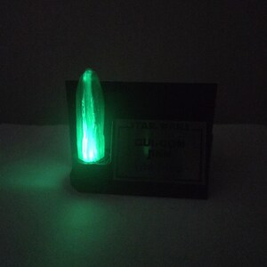 May include: A glowing green light saber prop, with the text "STAR WARS QUI-GON JINN" on a black base. The light saber emits a bright green glow, with a vertical blade shape.