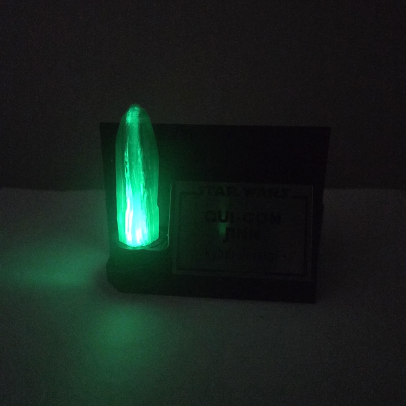 May include: A glowing green light saber prop, with the text "STAR WARS QUI-GON JINN" on a black base. The light saber emits a bright green glow, with a vertical blade shape.