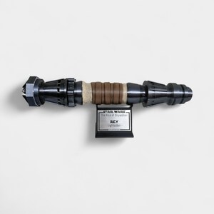 May include: A black and silver lightsaber prop from Star Wars: The Rise of Skywalker. The prop is labeled "Rey Lightsaber" and is mounted on a black stand.