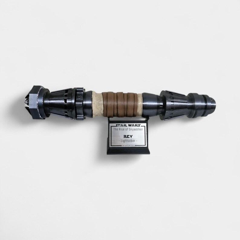 May include: A black and silver lightsaber prop from Star Wars: The Rise of Skywalker. The prop is labeled "Rey Lightsaber" and is mounted on a black stand.