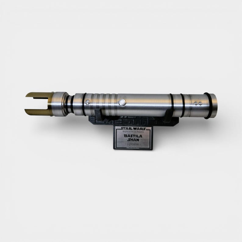 May include: A silver lightsaber with black accents and a gold blade emitter, mounted on a black stand. The stand features a plaque with the text "STAR WARS" and "BASTILA SHAN Lightsaber".