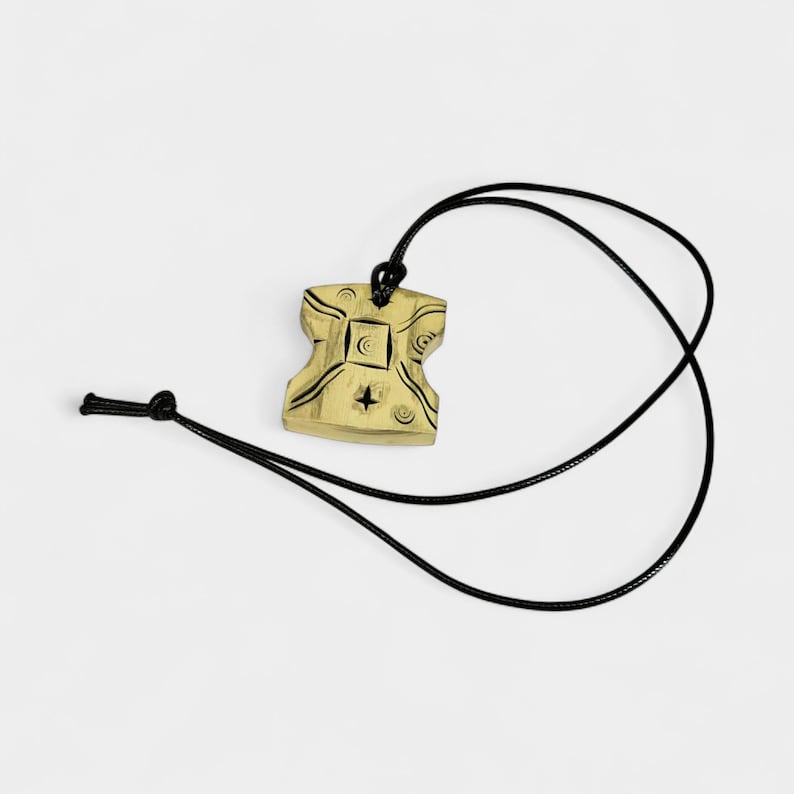 May include: A beige pendant with black etched designs, including a star and crescent moon, hangs from a black cord. The pendant has a unique, stylized shape. The cord is knotted at the top and bottom, creating an adjustable necklace.