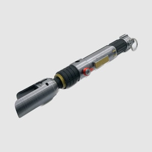 Pode incluir: Um sabre de luz prateado e preto com um bot&atilde;o vermelho e um detalhe dourado. O sabre de luz tem uma superf&iacute;cie texturizada e um pequeno la&ccedil;o na ponta.