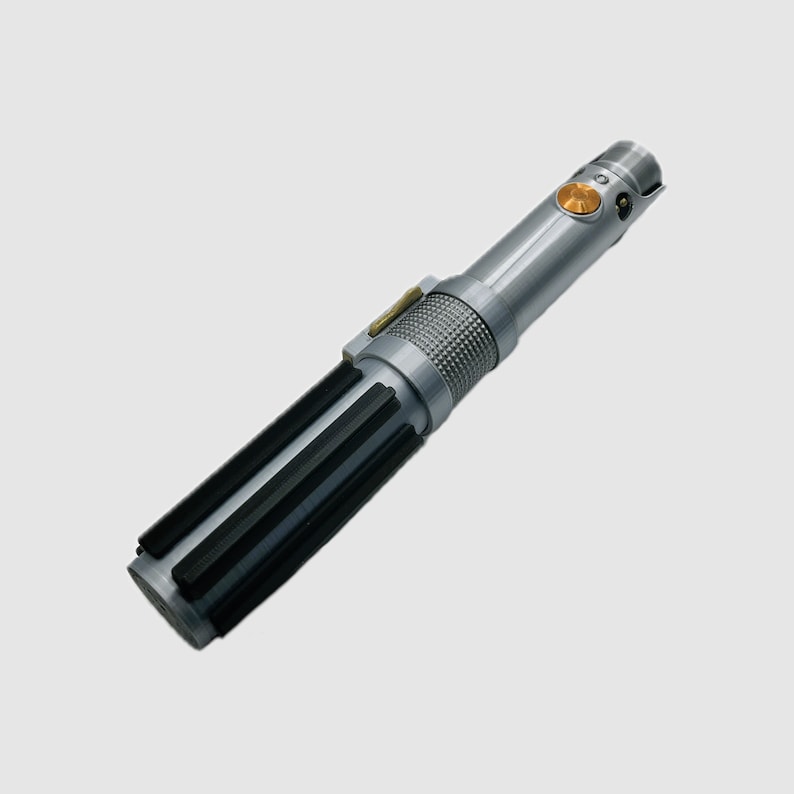 May include: A silver-toned cylindrical prop handle featuring a textured grip section with vertical black bars, a central knurled metallic band, and a circular gold-colored activation button positioned near the top emitter section.