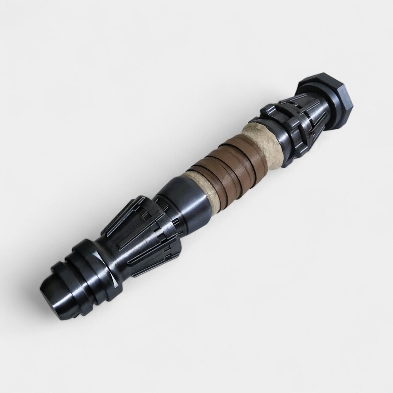 May include: A dark gray lightsaber with a brown leather wrap. The lightsaber has a textured, detailed design.