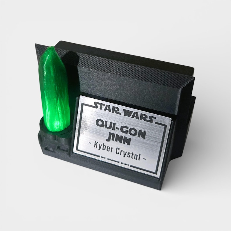 May include: A black display stand featuring a green Kyber Crystal and a silver plaque with the text "STAR WARS QUI-GON JINN - Kyber Crystal". The stand is a Star Wars collectible.