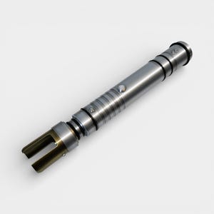 May include: A silver lightsaber with a metallic finish. The hilt features a button and several black rings. The emitter is a gold color with two prongs. The lightsaber is a prop or toy.