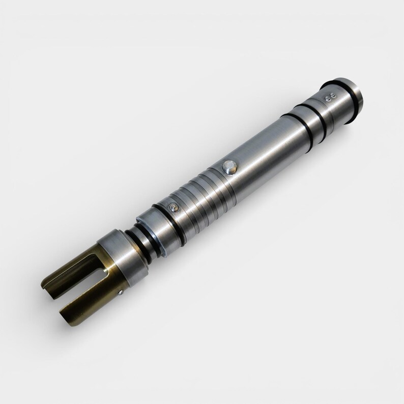May include: A silver lightsaber with a metallic finish. The hilt features a button and several black rings. The emitter is a gold color with two prongs. The lightsaber is a prop or toy.