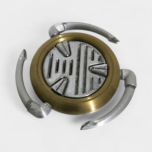 Op de afbeelding: Een metalen fidget spinner met drie gebogen armen. Het ronde middenstuk heeft een zilverkleurig patroon in een messingkleurige ring. De drie armen zijn zilverkleurig.