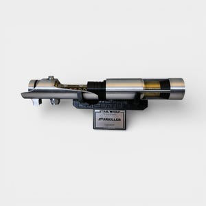 May include: A silver and black Starkiller lightsaber prop from Star Wars, displayed on a black stand. The lightsaber has a detailed design with a metallic finish and a clear section revealing internal components. The stand features the Star Wars logo.