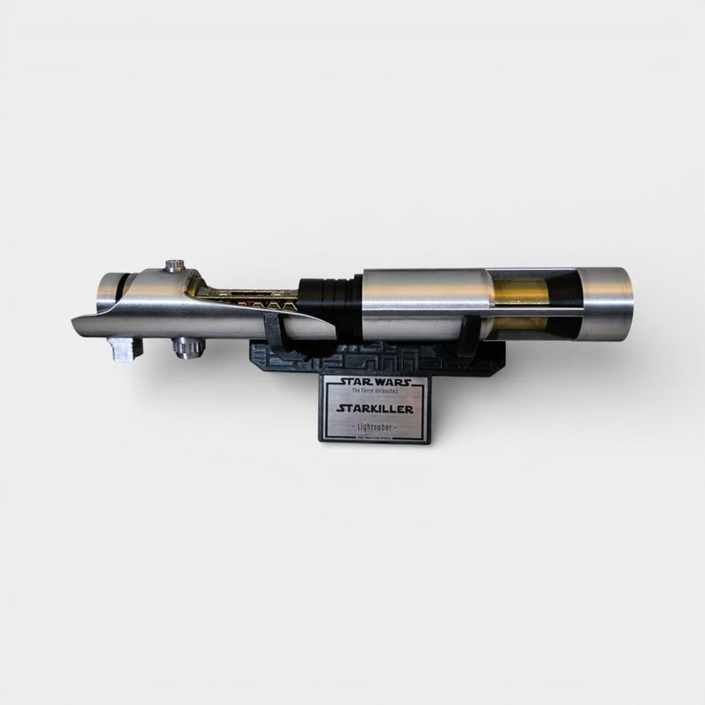 May include: A silver and black Starkiller lightsaber prop from Star Wars, displayed on a black stand. The lightsaber has a detailed design with a metallic finish and a clear section revealing internal components. The stand features the Star Wars logo.