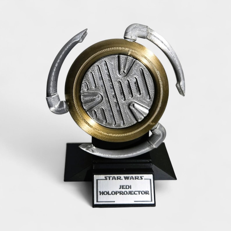 Op de afbeelding: Een replica van een Star Wars Jedi Holoprojector. Het apparaat heeft een zilver en goud cirkelvormig ontwerp met drie gebogen armen. Het staat op een zwarte basis met een naamplaatje met de tekst "Star Wars Jedi Holoprojector."