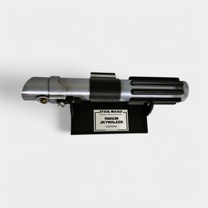 May include: A black lightsaber prop from the Star Wars movie franchise. The lightsaber is mounted on a black stand with a silver plaque that reads "STAR WARS Episode II Attack of the Clones ANAKIN SKYWALKER - Lightsaber - "