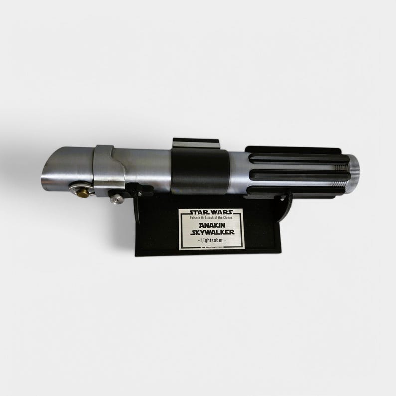 May include: A black lightsaber prop from the Star Wars movie franchise. The lightsaber is mounted on a black stand with a silver plaque that reads "STAR WARS Episode II Attack of the Clones ANAKIN SKYWALKER - Lightsaber - "