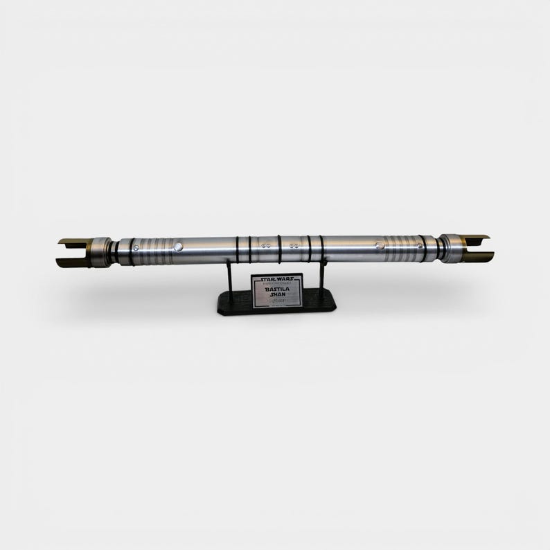 May include: A silver lightsaber prop, displayed horizontally on a black stand. The lightsaber has detailed metallic accents and a blade emitter on each end. The stand features a label that reads "STAR WARS BASTILA SHAN".