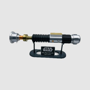 May include: A replica Obi-Wan Kenobi lightsaber hilt featuring a silver, black, and gold metallic finish, mounted on a black display stand. The stand includes a plaque with the text STAR WARS Obi-Wan Kenobi Lightsaber in white lettering.