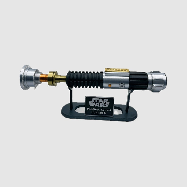 May include: A replica Obi-Wan Kenobi lightsaber hilt featuring a silver, black, and gold metallic finish, mounted on a black display stand. The stand includes a plaque with the text STAR WARS Obi-Wan Kenobi Lightsaber in white lettering.