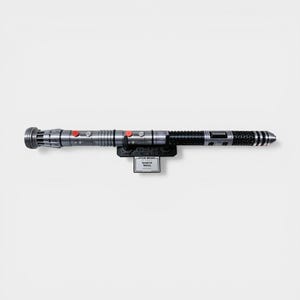 Puede incluir: Un sable de luz plateado y negro, montado en un soporte negro. El sable de luz tiene botones rojos y un dise&ntilde;o detallado. El soporte presenta el logotipo de Star Wars y el nombre "Darth Maul".