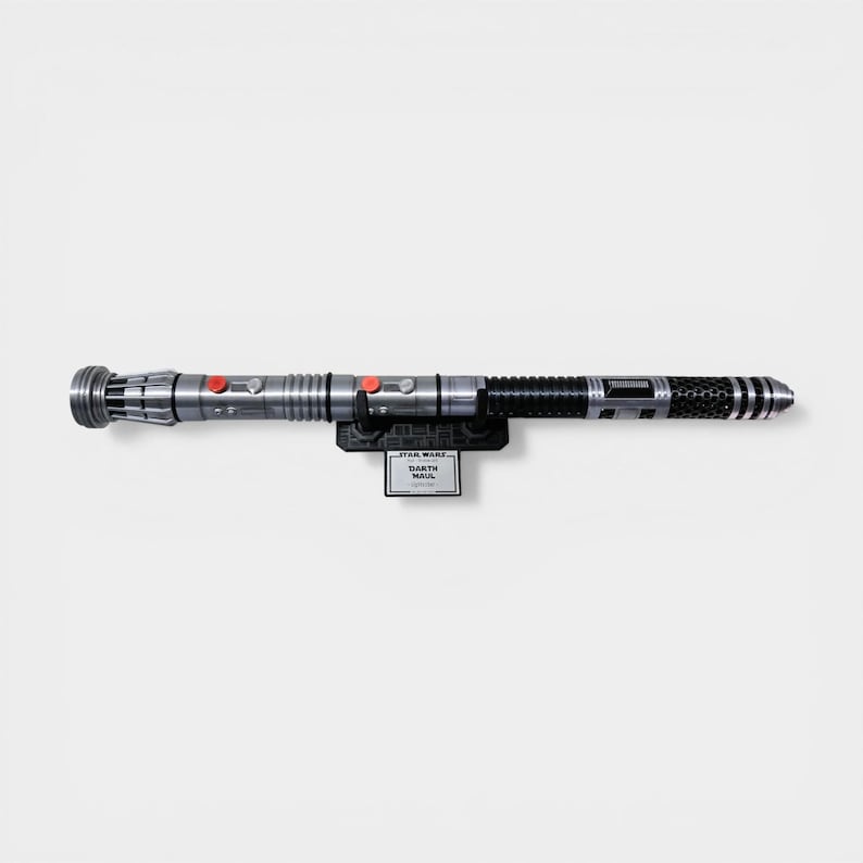 Puede incluir: Un sable de luz plateado y negro, montado en un soporte negro. El sable de luz tiene botones rojos y un dise&ntilde;o detallado. El soporte presenta el logotipo de Star Wars y el nombre "Darth Maul".