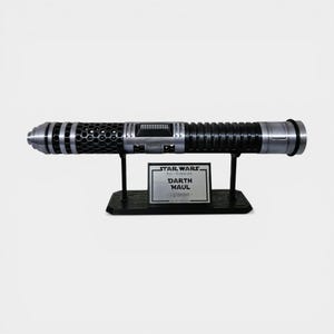 Puede incluir: Una r&eacute;plica del sable de luz Darth Maul de Star Wars, negro y plateado, exhibida en un soporte negro. El sable de luz presenta detalles intrincados y un acabado met&aacute;lico. Una placa plateada en el soporte dice "Star Wars Darth Maul Lightsaber."
