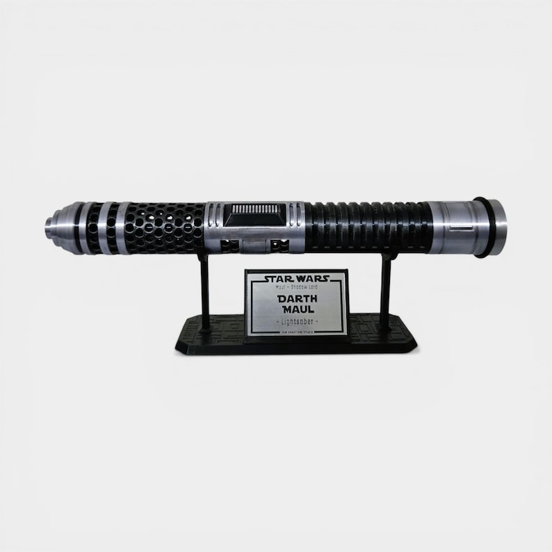 Puede incluir: Una r&eacute;plica del sable de luz Darth Maul de Star Wars, negro y plateado, exhibida en un soporte negro. El sable de luz presenta detalles intrincados y un acabado met&aacute;lico. Una placa plateada en el soporte dice "Star Wars Darth Maul Lightsaber."