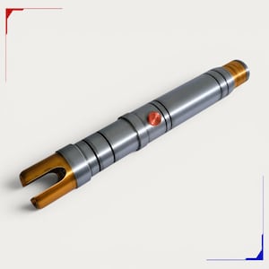 May include: A cylindrical metal prop with a matte grey finish, featuring a gold-toned emitter at one end and a matching gold-toned pommel at the other. A small, circular, copper-coloured activation button is positioned on the central handle section.