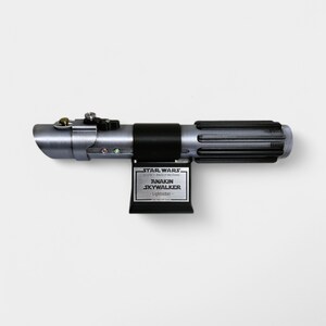 May include: A silver and black lightsaber with a black plaque that reads "Star Wars Episode II: Attack of the Clones Anakin Skywalker Lightsaber".