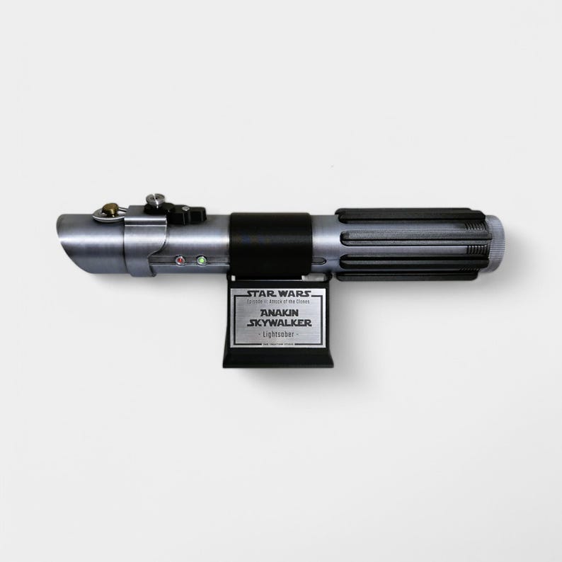 May include: A silver and black lightsaber with a black plaque that reads "Star Wars Episode II: Attack of the Clones Anakin Skywalker Lightsaber".