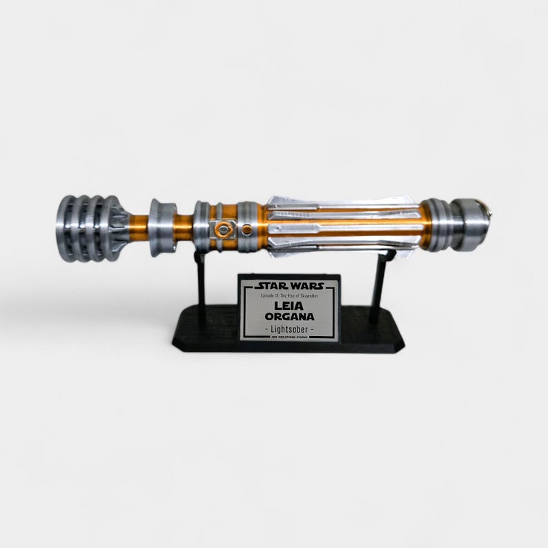 Puede incluir: R&eacute;plica de sable de luz plateada y dorada, presentada sobre un soporte negro. El sable de luz tiene detalles intrincados y est&aacute; etiquetado como "Leia Organa - Lightsaber" en una peque&ntilde;a placa. El logotipo de Star Wars tambi&eacute;n es visible.