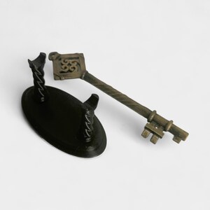 May include: An antique-style key with a decorative handle and a twisted shaft, next to a black oval stand with two curved supports. The key has a bronze finish and intricate detailing.