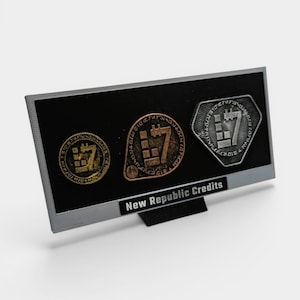 May include: A display of three New Republic Credits coins. The coins are in gold, copper, and silver tones, each featuring a stylized "7" design. The coins are set against a black background within a gray frame, with the text "New Republic Credits" below.